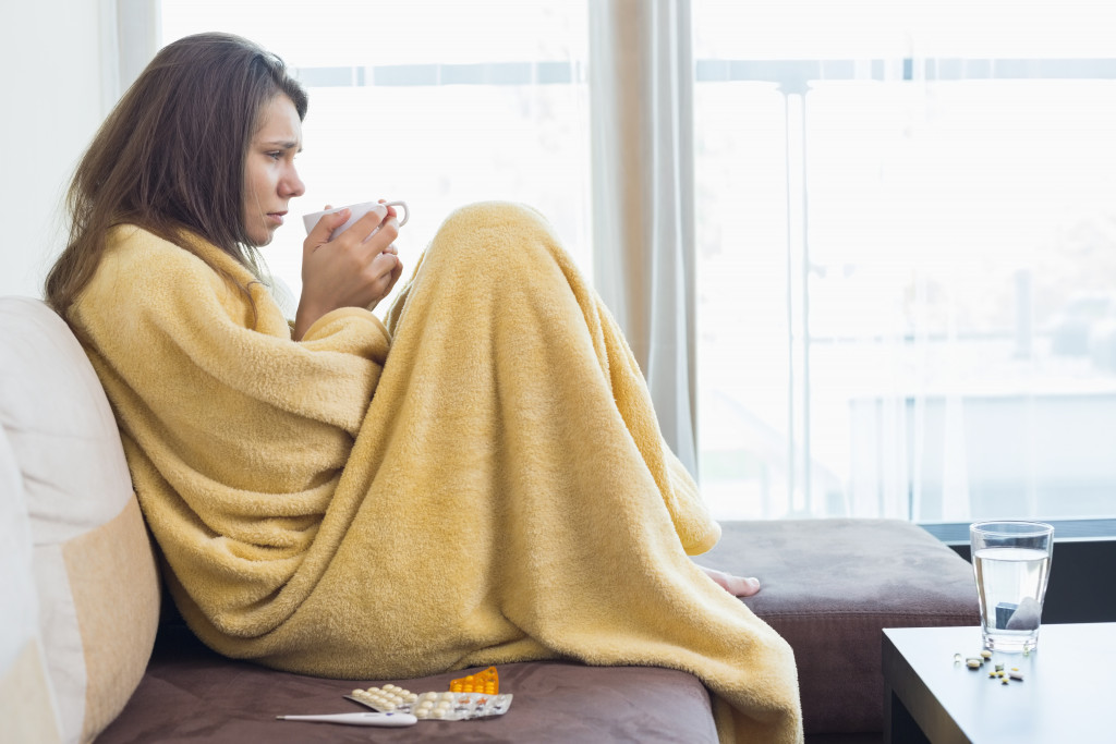 Sick young woman sitting on a sofa while drinking coffee and wrapped in a blanket