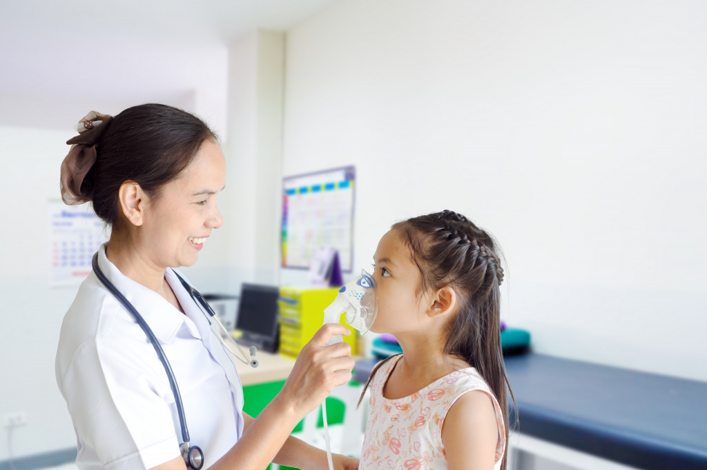 Lady doctor holding an oxygen mask as a little girl breathes it in inside a clinic.