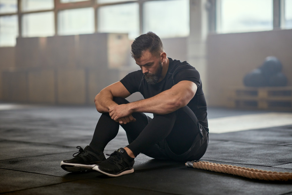 Fit young man in sportswear sitting on a gym floor and looking exhausted after a battle rope workout session